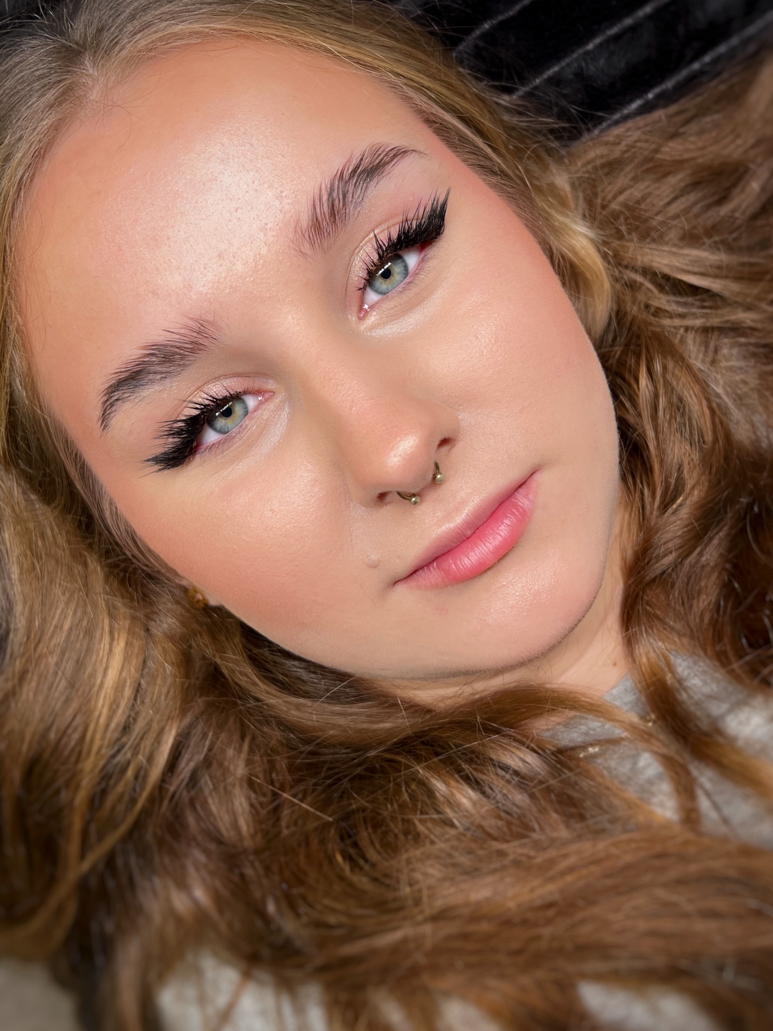 Portret młodej kobiety z profesjonalnie przedłużonymi rzęsami w stylu anime, delikatnym różem na policzkach i kolczykiem w nosie typu septum, leżącej na czarnym tle.