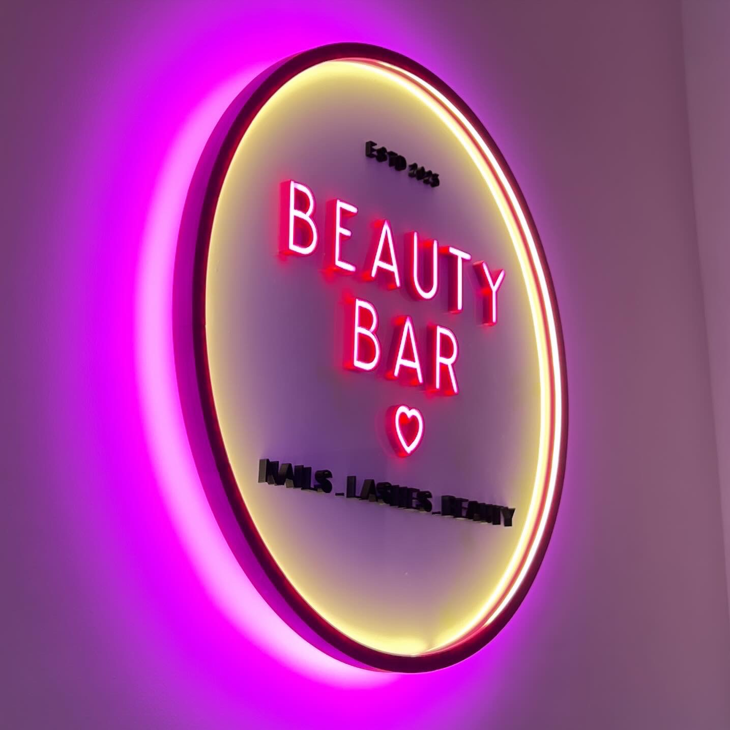 Neonowy szyld 'Beauty Bar' z napisem 'Nails Lashes Beauty' u dołu, oświetlony różowym i żółtym światłem, zamontowany na ścianie.