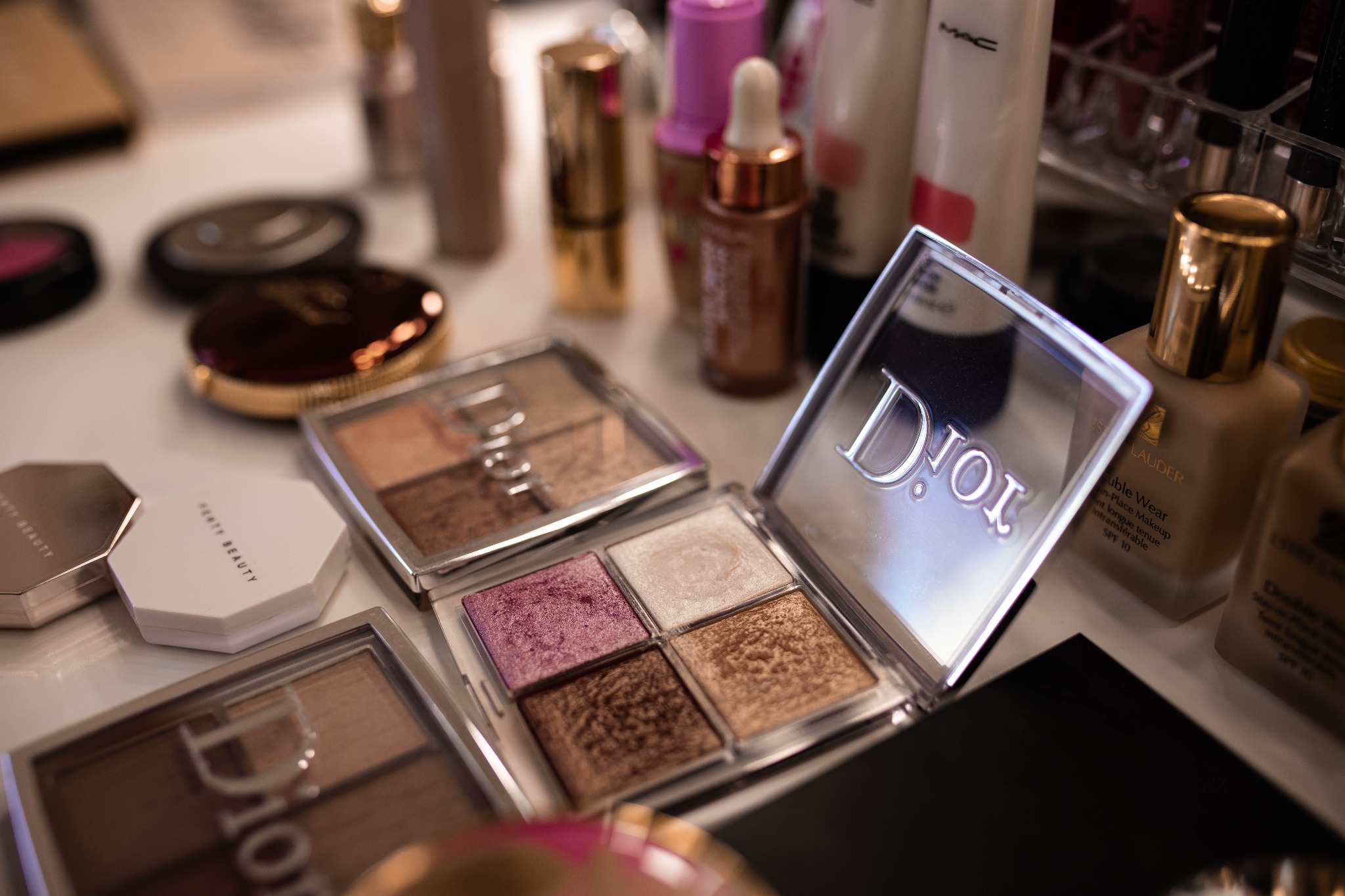 Rozłożone palety cieni do powiek marek Dior i Fenty Beauty oraz podkłady Estee Lauder i MAC na białym stole w salonie kosmetycznym.