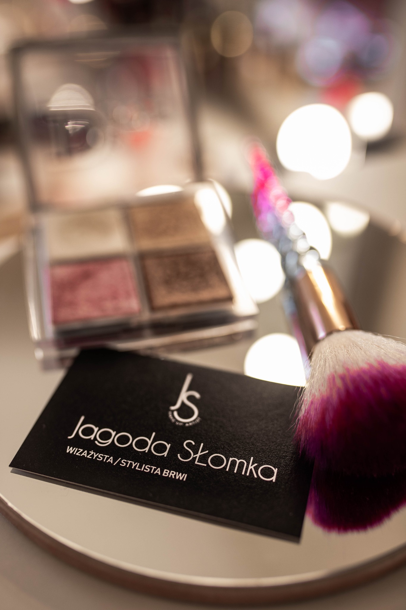 Paleta cieni do powiek, wizytówka 'Jagoda Słomka Wizażysta/Stylista Brwi' oraz pędzel do makijażu z różowym włosiem na okrągłym lustrze.