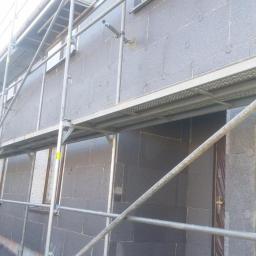 Bakis - Ocieplanie elewacji budynku styropianem grafitowym, widoczne rusztowanie i fragment ogrodu z zielonymi krzewami.