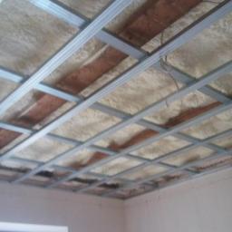 Bakis - Sufit w trakcie izolacji pianą natryskową, widoczna metalowa konstrukcja podwieszana, częściowo zakryta pianą, odsłonięte fragmenty starego stropu.