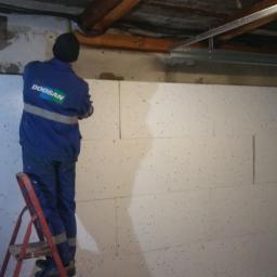 Bakis - Pracownik w niebieskim kombinezonie z logo Doosan na drabinie montuje płyty izolacyjne na ścianie w garażu, widoczne rury pod sufitem i metalowa konstrukcja nad bramą garażową.