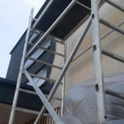 Bakis - Przygotowania do prac na elewacji budynku: rusztowanie, folia ochronna i świeżo pomalowany fragment ściany na ciemny kolor.