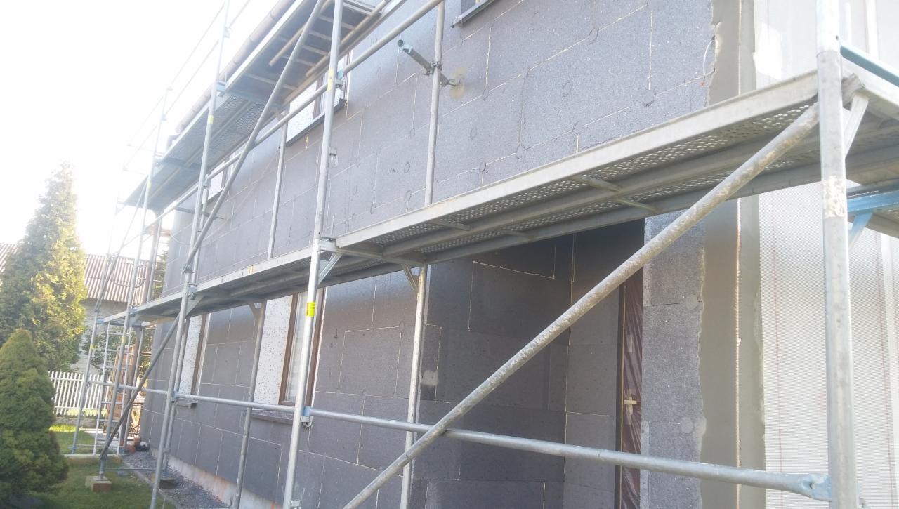 Ocieplanie elewacji budynku styropianem grafitowym, widoczne rusztowanie i fragment ogrodu z zielonymi krzewami.