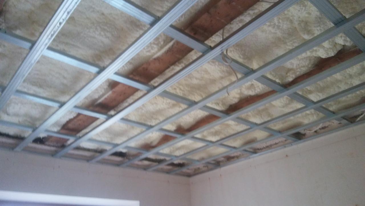 Sufit w trakcie izolacji pianą natryskową, widoczna metalowa konstrukcja podwieszana, częściowo zakryta pianą, odsłonięte fragmenty starego stropu.