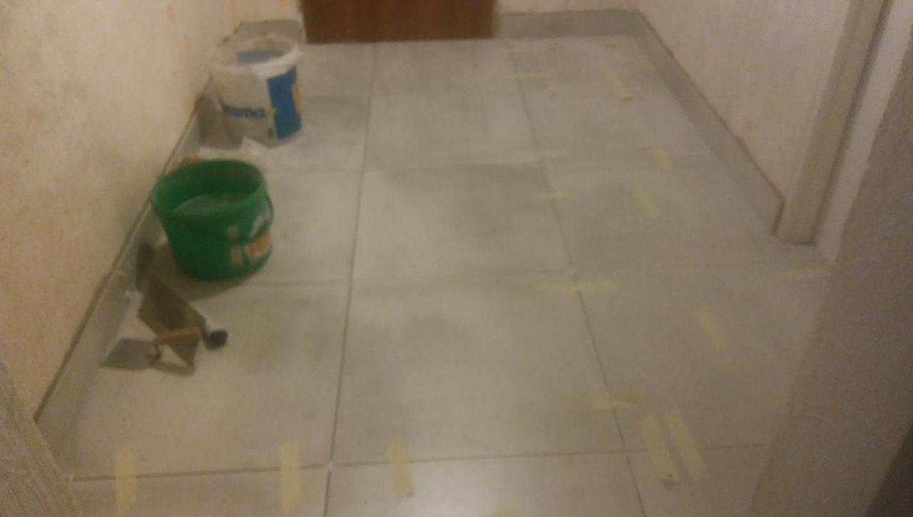 Przygotowanie do fugowania szarych płytek podłogowych w korytarzu, widoczne taśmy maskujące, puste wiaderka i kielnie.