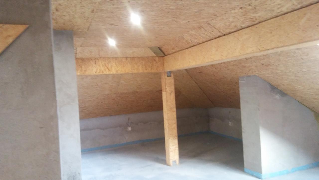 Poddasze w trakcie adaptacji: sufit wykończony płytami OSB, widoczne drewniane belki konstrukcyjne i słupy, ściany surowe, przygotowane do dalszych prac wykończeniowych.