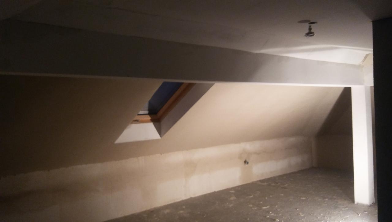 Poddasze w trakcie prac wykończeniowych; widoczna ściana ze śladami po szpachlowaniu, okno dachowe w drewnianej ramie i surowy betonowy strop.