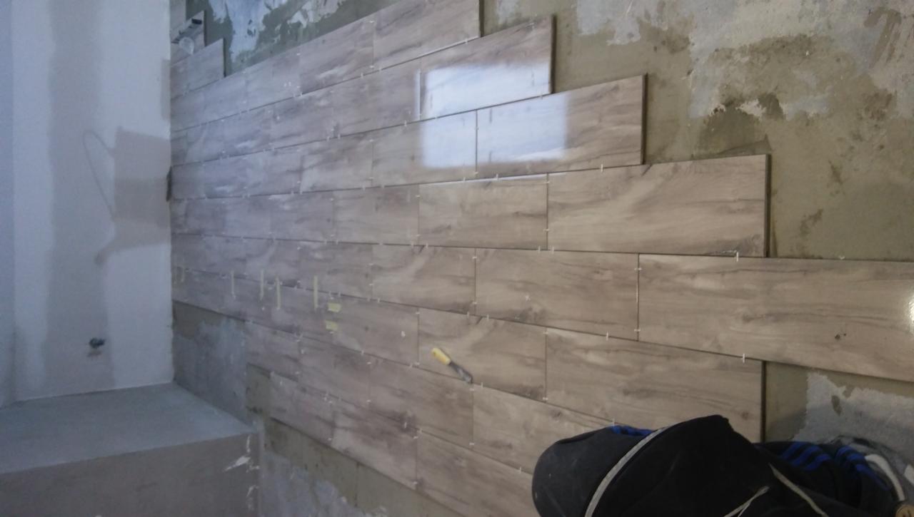 Układanie podłużnych płytek imitujących drewno na ścianie w trakcie remontu, widoczne krzyżyki dystansowe i fragment ściany bez płytek.