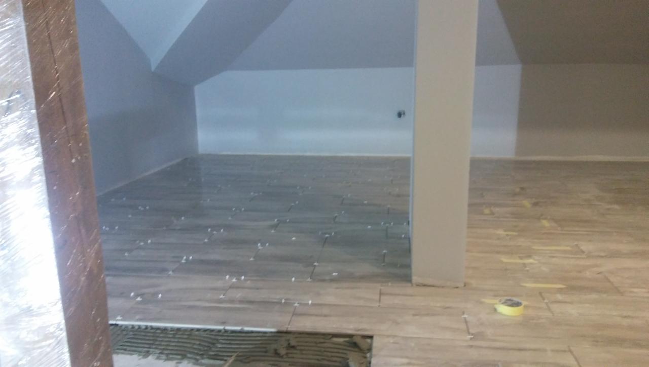 Poddasze w trakcie układania podłogi z płytek imitujących drewno; widoczne fugi, częściowo ułożona podłoga oraz taśma malarska.