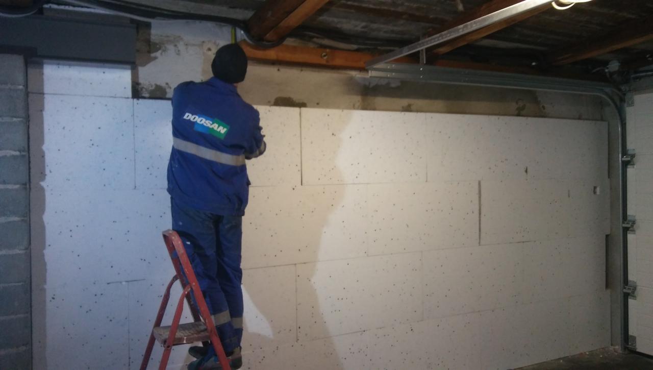 Pracownik w niebieskim kombinezonie z logo Doosan na drabinie montuje płyty izolacyjne na ścianie w garażu, widoczne rury pod sufitem i metalowa konstrukcja nad bramą garażową.