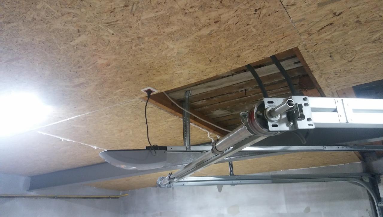 Montaż automatyki bramy garażowej: widoczny silnik, szyna prowadząca i sprężyna na tle surowego sufitu z płyty OSB z otworem inspekcyjnym.