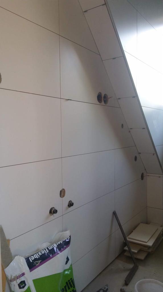 Ściana w trakcie układania białych płytek w łazience na poddaszu, widoczne otwory na instalacje i worek z klejem do płytek.