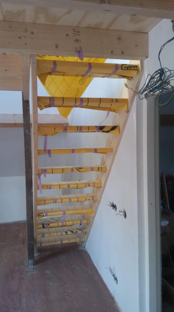 Drewniane schody w trakcie zabezpieczania żółtą folią ochronną, przymocowaną fioletową taśmą, widoczne niedokończone instalacje elektryczne w ścianie.