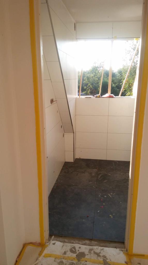 Wnętrze łazienki w trakcie prac wykończeniowych, widoczne białe płytki na ścianach i ciemna podłoga z płytek, zabezpieczone taśmą malarską krawędzie ścian i podłogi, okno z widokiem na zieleń.