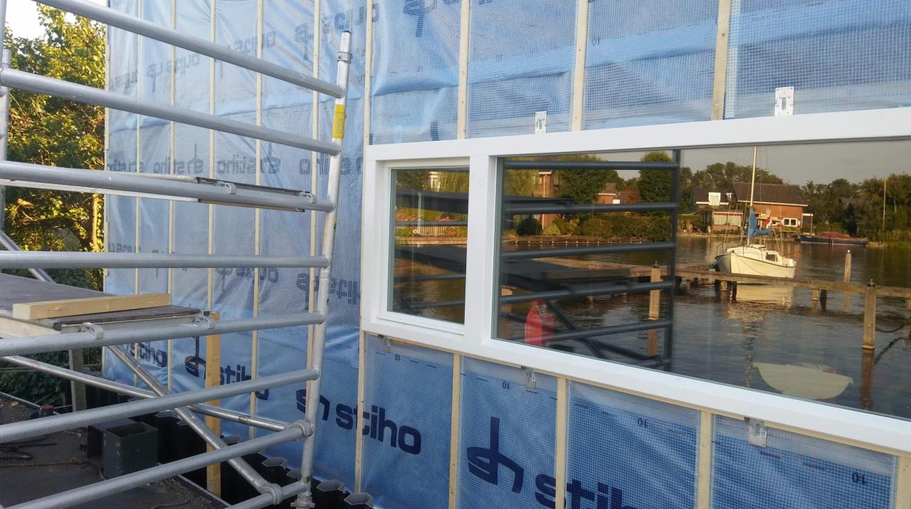 Montaż okna z białymi ramami w budynku w trakcie budowy, widoczne rusztowanie i folia ochronna na ścianie, w szybie okna odbija się krajobraz z jeziorem i łódką.