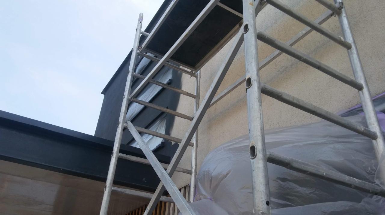 Przygotowania do prac na elewacji budynku: rusztowanie, folia ochronna i świeżo pomalowany fragment ściany na ciemny kolor.