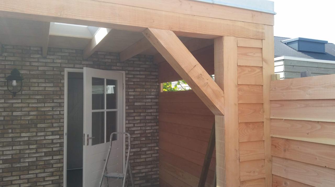 Drewniana konstrukcja zadaszenia nad wejściem, połączona z ceglaną ścianą budynku i drewnianym ogrodzeniem, białe drzwi częściowo otwarte, drabina oparta o ścianę.