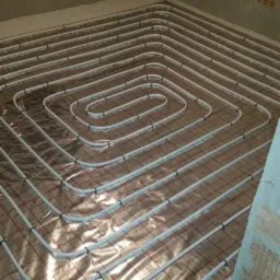 Białe rury ogrzewania podłogowego ułożone spiralnie na folii izolacyjnej z czerwoną siatką, widok z góry w trakcie montażu.