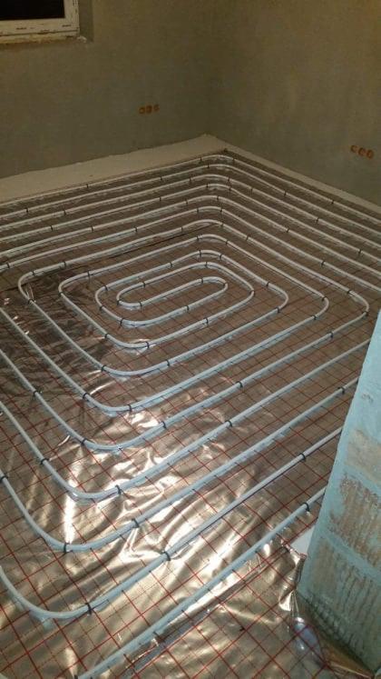 Białe rury ogrzewania podłogowego ułożone spiralnie na folii izolacyjnej z czerwoną siatką, widok z góry w trakcie montażu.