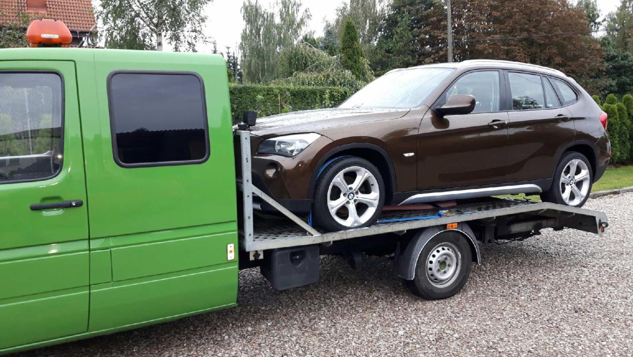 Brązowe BMW X1 na zielonej lawecie w Warszawie, zabezpieczone niebieskimi pasami transportowymi.