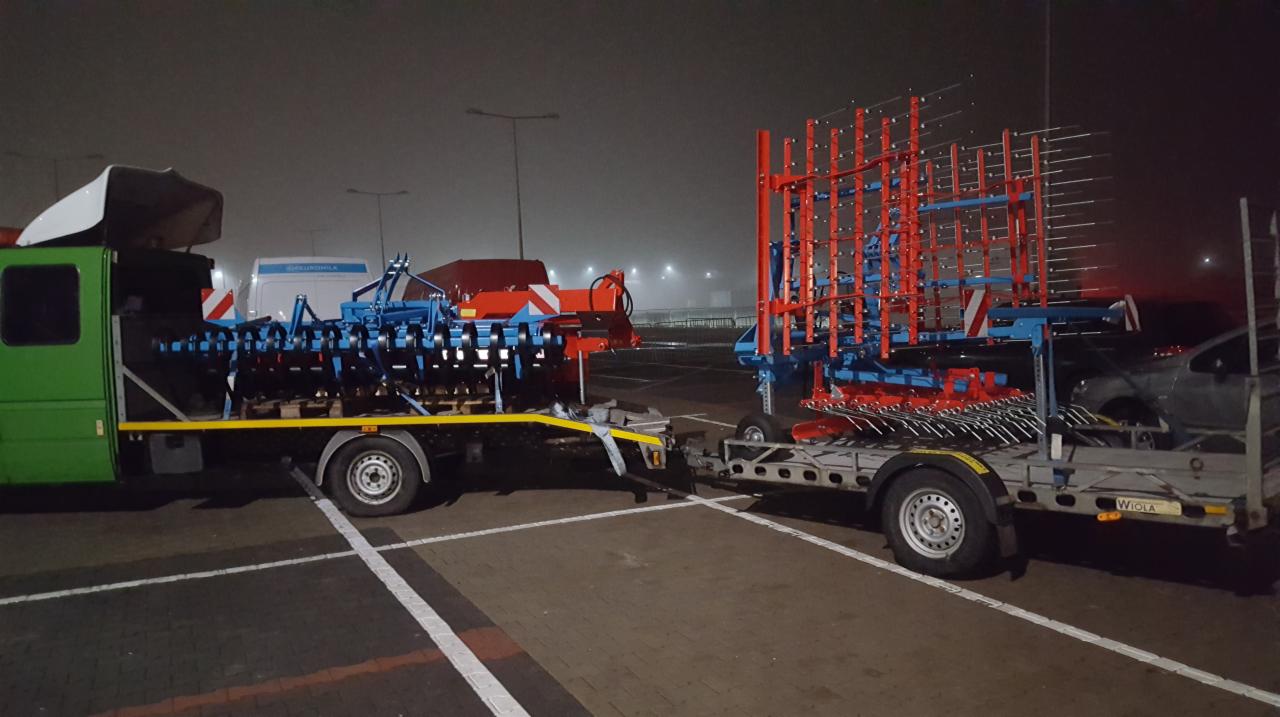 Zielony bus z białą nadbudówką i przyczepą przewozi niebiesko-czerwone maszyny rolnicze na parkingu w pochmurny dzień.