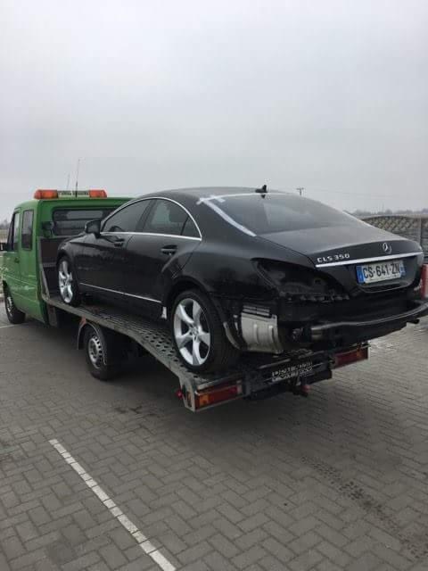 Czarny uszkodzony Mercedes CLS 350 na zielonej lawecie z pomarańczowymi światłami ostrzegawczymi, widok z tyłu, tablica rejestracyjna CS-641-ZN.