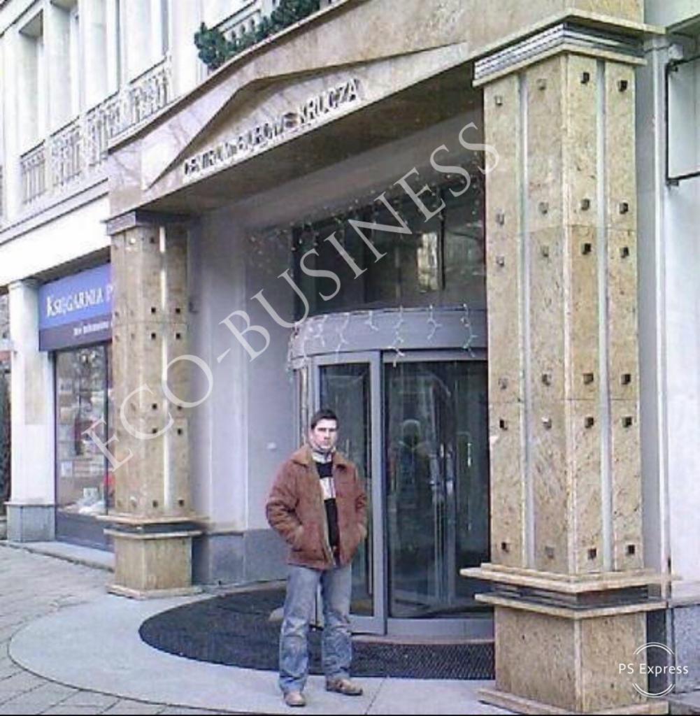 Wejście do budynku z obrotowymi drzwiami, ozdobione kamiennymi kolumnami z regularnymi wgłębieniami, na tle fasady z balkonem i szyldem, z mężczyzną stojącym przed wejściem.
