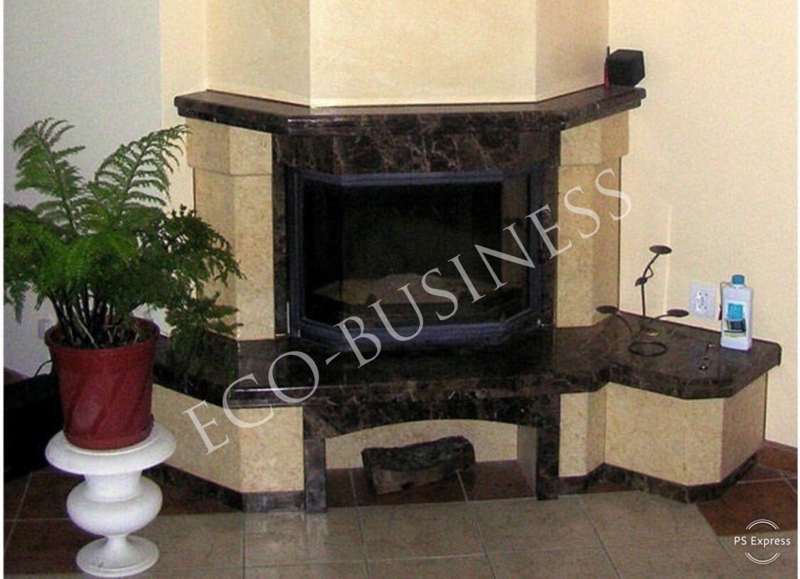 Narożny kominek z obudową z jasnego i ciemnego marmuru, z szybą i dekoracyjnym drewnem w podstawie, obok doniczka z paprotką na białym postumencie i dekoracje na blacie.