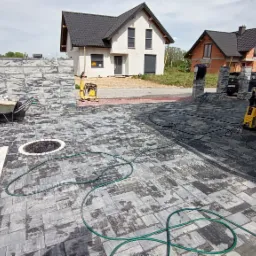 Ułożona kostka brukowa w odcieniach szarości z białymi obramowaniami, widoczna taczka z materiałem, zagęszczarka wibracyjna i myjka ciśnieniowa, dom w tle.