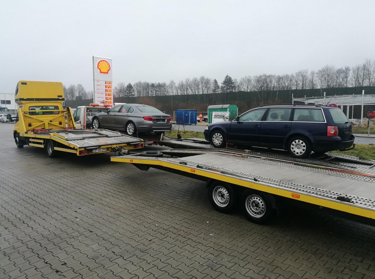 Żółta laweta z dwoma samochodami osobowymi na platformach, zaparkowana przy stacji benzynowej Shell w pochmurny dzień.