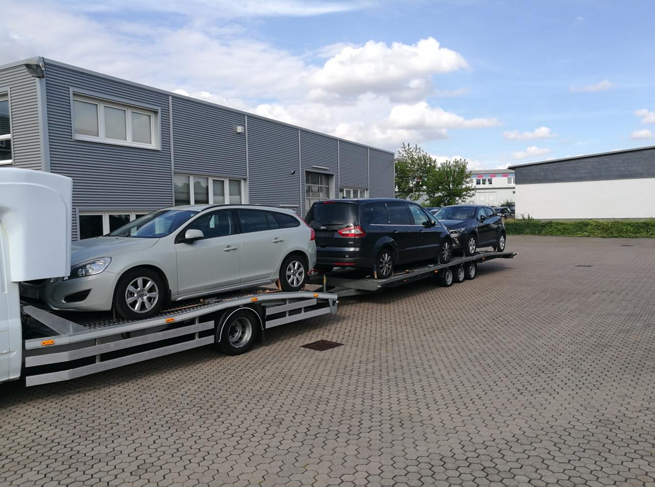 Laweta z trzema samochodami osobowymi: srebrne Volvo, czarny Ford Galaxy i czarny Nissan. Transport aut na tle szarego budynku i błękitnego nieba.