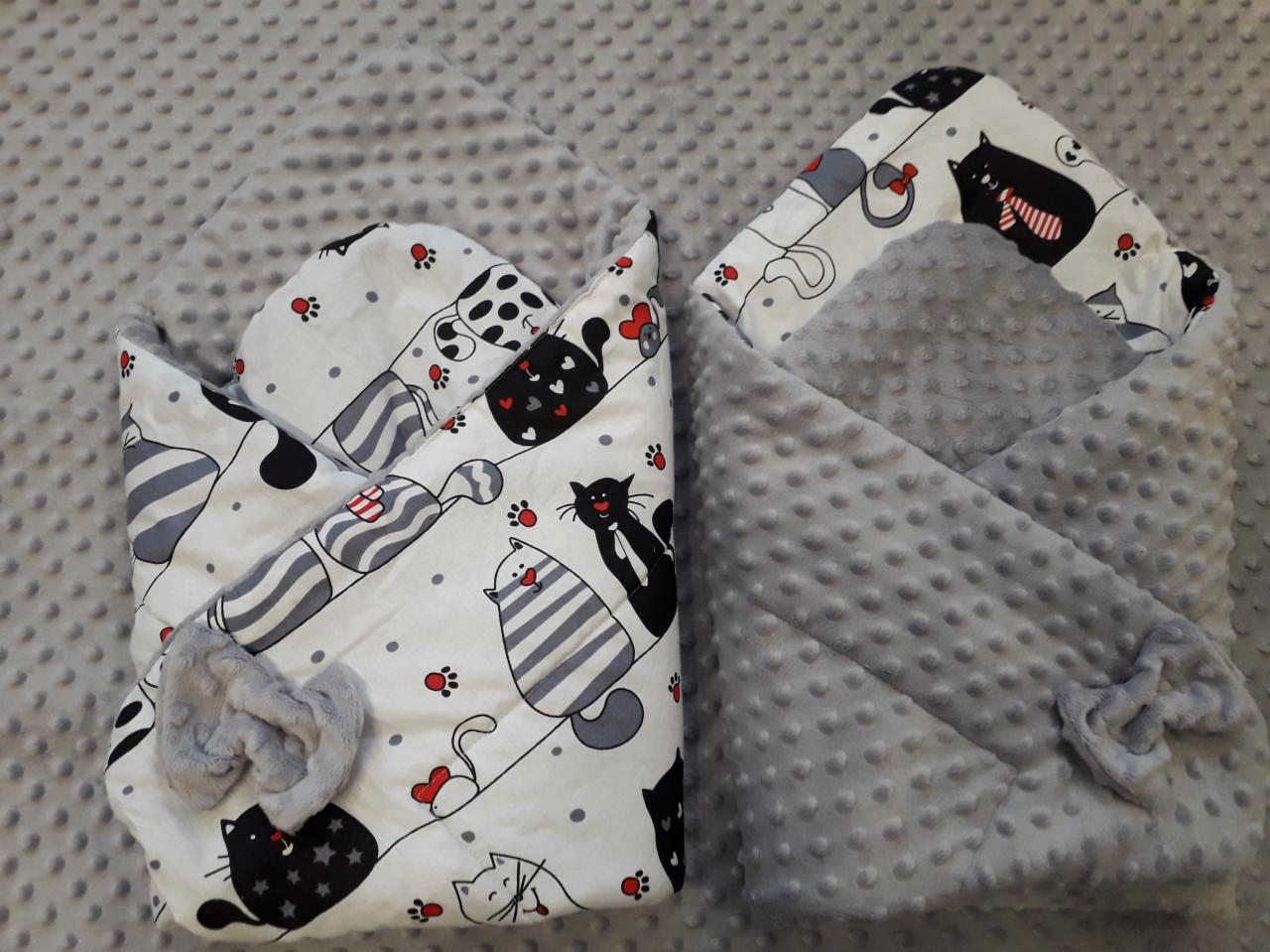 Dwa otulacze niemowlęce: jeden biały w czarne koty z szarą kokardką, drugi szary z fakturą w kropki i szarą kokardką, leżące na szarym tle.
