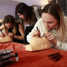 Kurs przedłużania rzęs: Uczestniczki ćwiczą aplikację rzęs na manekinie głowy z bliska, widoczne pęsety i opakowania rzęs na czerwonym stole.