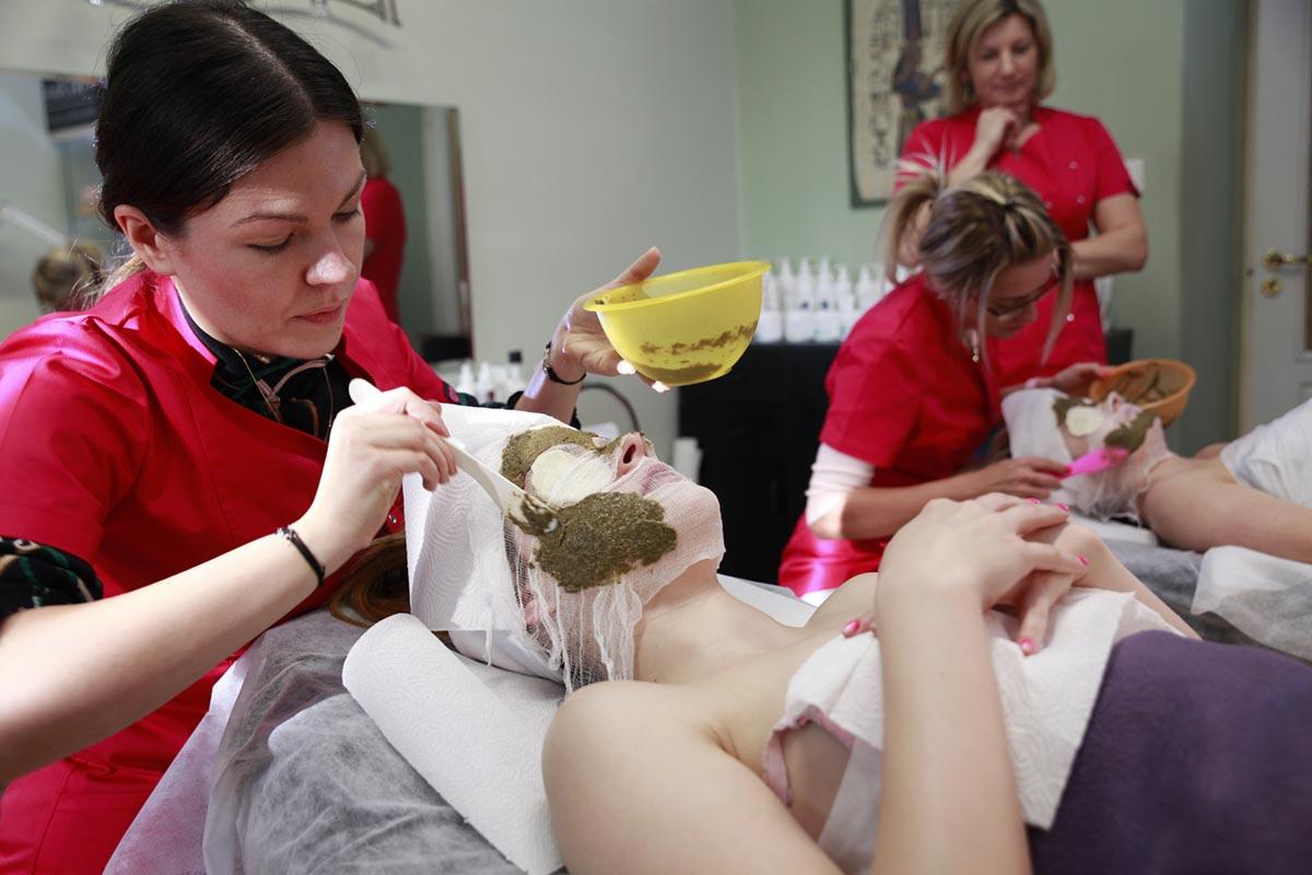 Kosmetolog nakłada zieloną maskę na twarz klientki leżącej na leżance w salonie kosmetycznym, widoczne inne klientki i pracownice w tle.