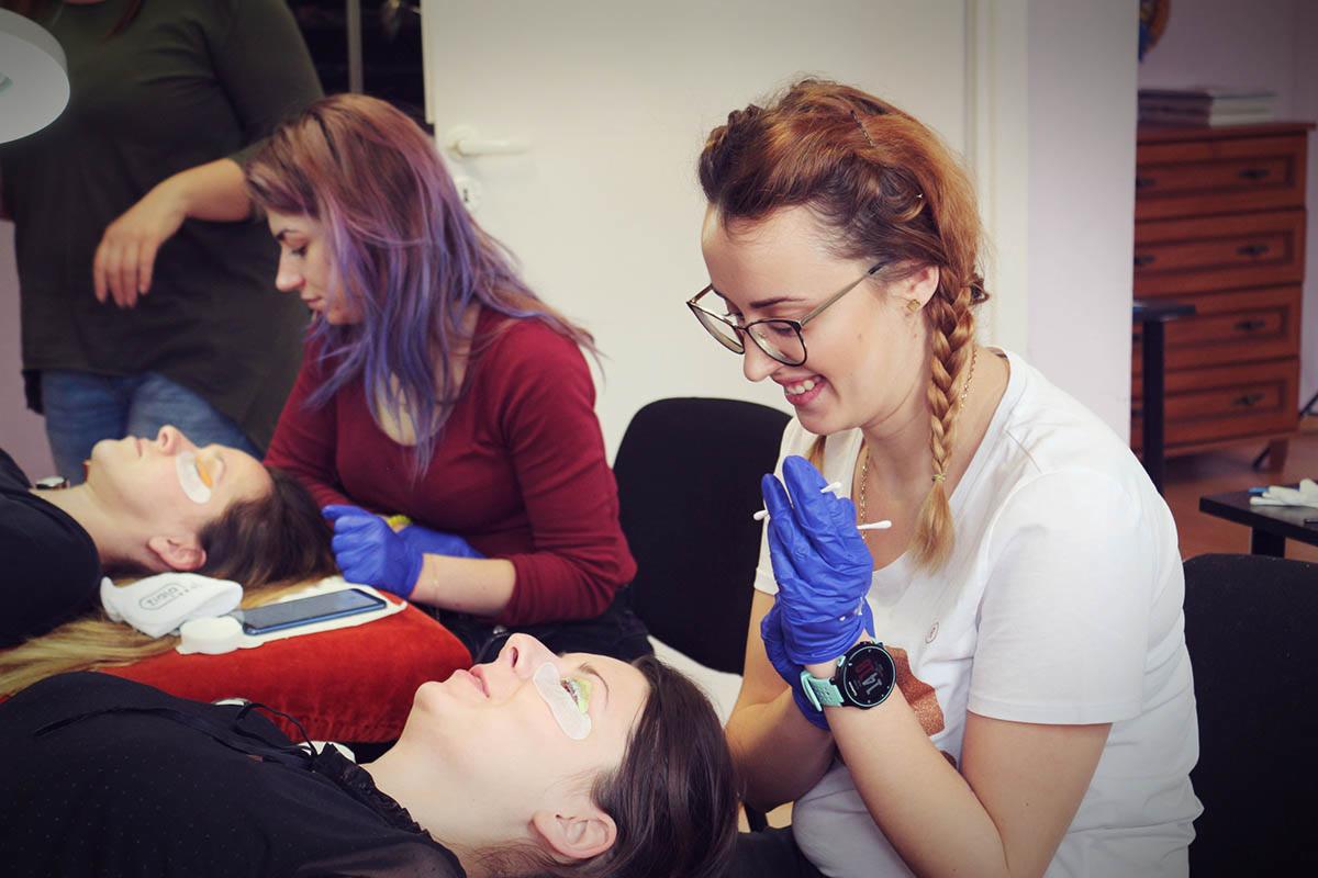 Stylistka rzęs w niebieskich rękawiczkach uśmiecha się podczas aplikacji rzęs klientce leżącej na czerwonej poduszce; obok druga stylistka z fioletowymi włosami pracuje nad drugą klientką.