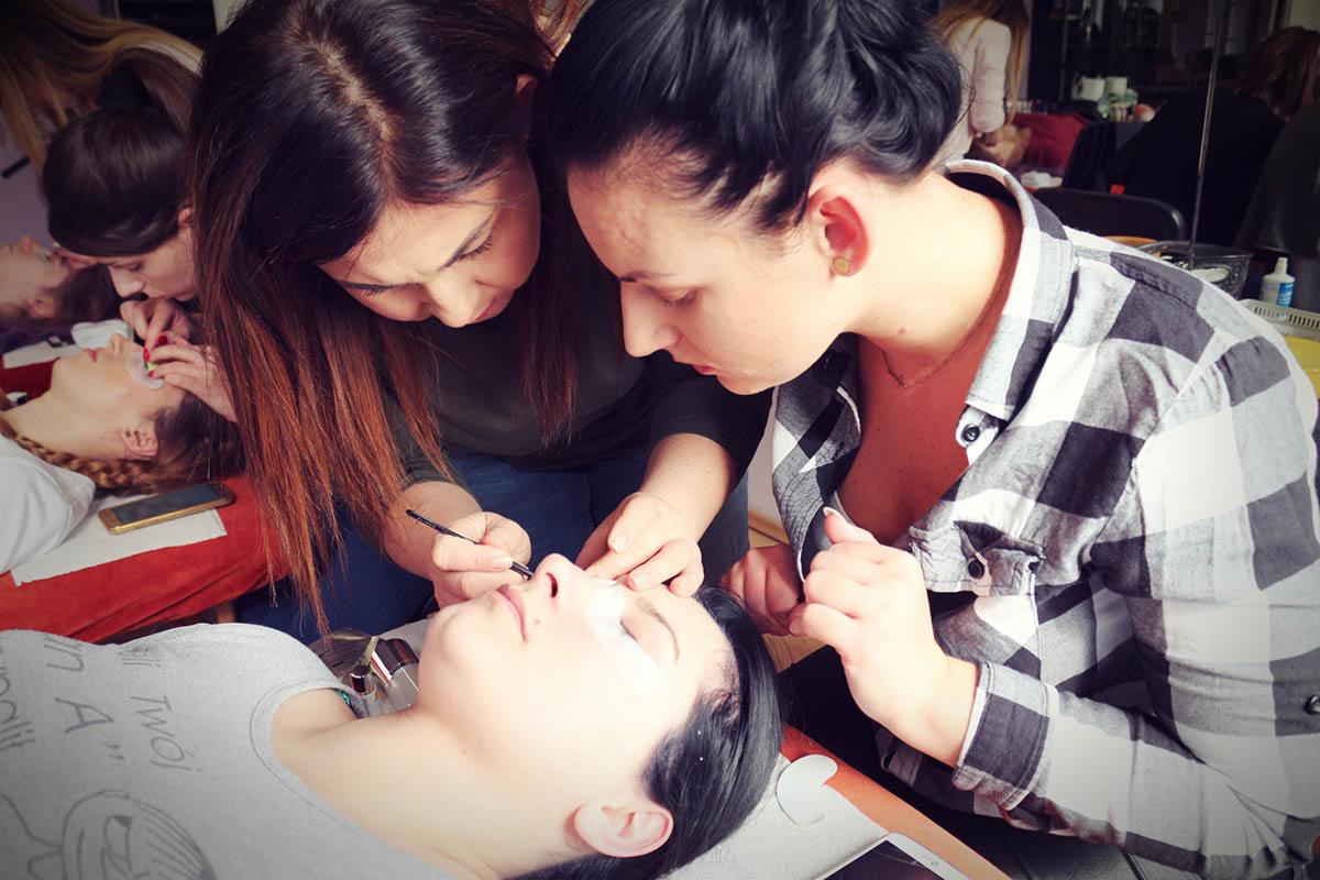 Kurs przedłużania rzęs: kursantka w koszuli w kratę aplikuje rzęsy pod okiem instruktora, w tle inne stanowiska pracy.