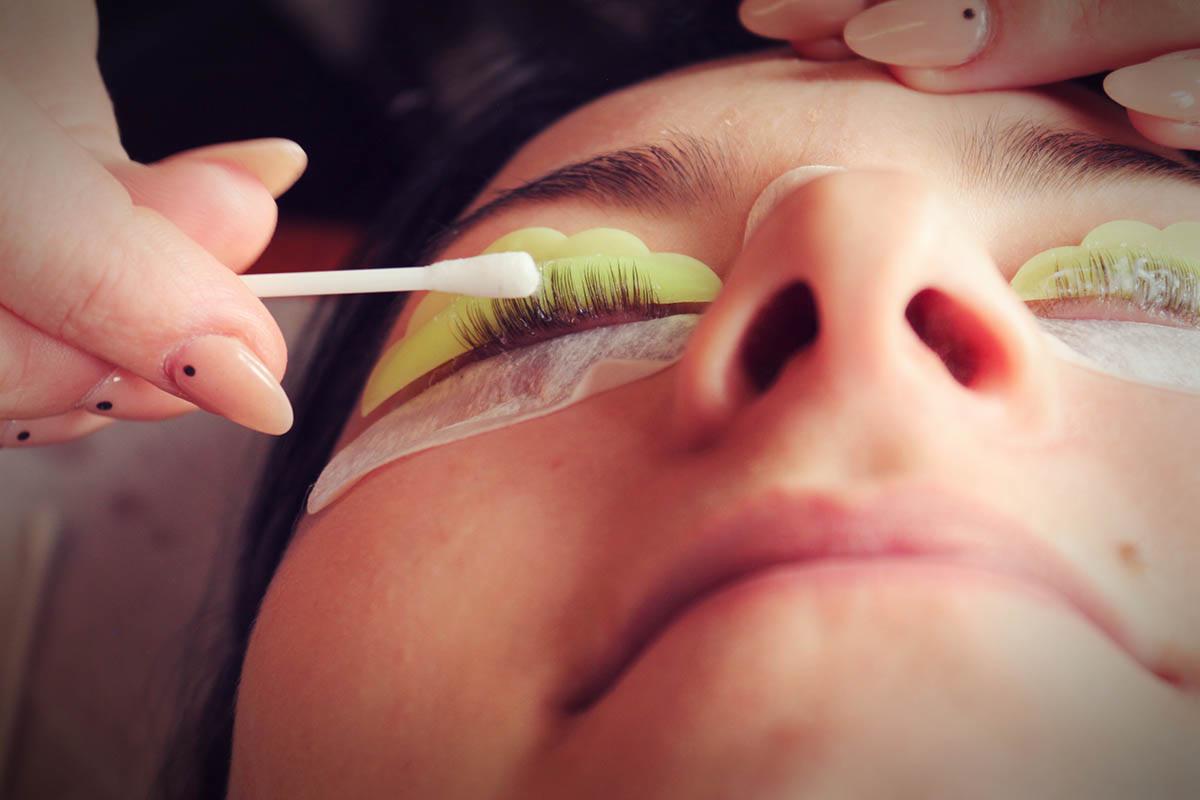 Zabieg liftingu rzęs: rzęsy klientki na wałeczku silikonowym, kosmetyczka aplikuje preparat wacikiem, widoczne dłonie kosmetyczki z pomalowanymi paznokciami.