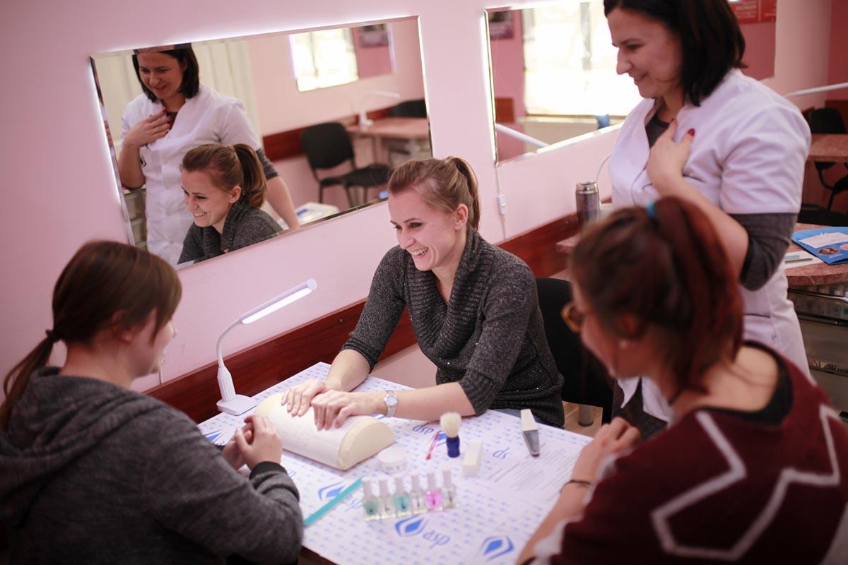 Kurs manicure: Uczestniczka uśmiechnięta, ćwiczy na sztucznej dłoni pod okiem instruktorki w białym uniformie, odbijającej się w lustrze, otoczona przez inne kursantki.