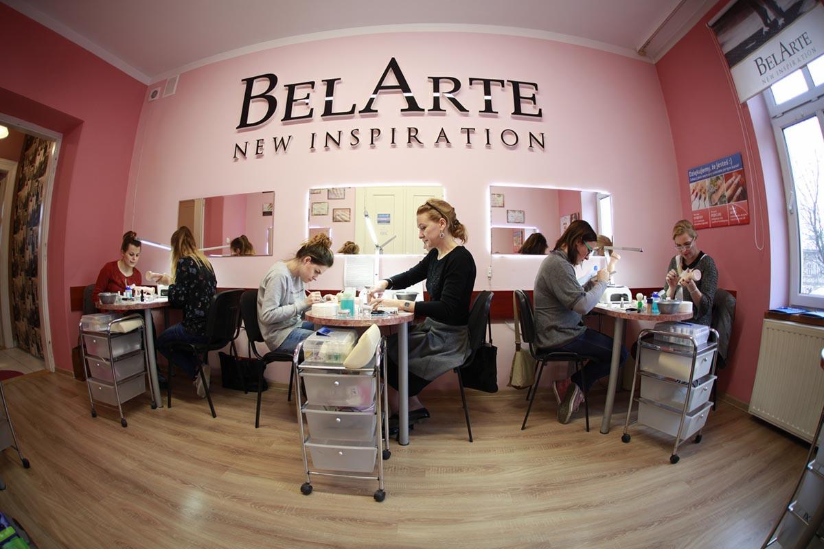Wnętrze salonu kosmetycznego z czterema stanowiskami do manicure, kobiety wykonują zabiegi, widoczne lampki, lakiery i akcesoria, na ścianie logo 'BelArte New Inspiration'.