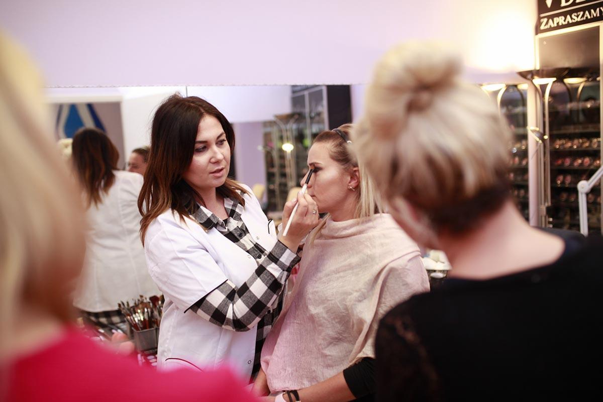 Wizażystka w koszuli w kratę aplikuje makijaż pędzlem na powiekę klientki, w tle lustro i kosmetyki na półkach.