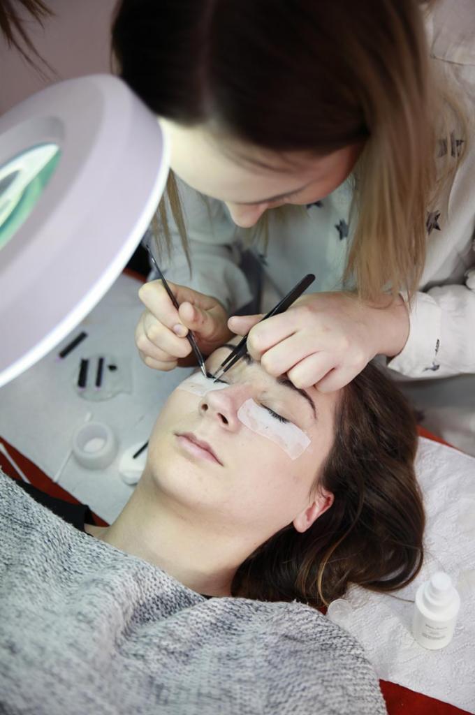 Przedłużanie rzęs: stylistka aplikuje sztuczne rzęsy pęsetą, klientka leży z zamkniętymi oczami pod lampą powiększającą, zabezpieczona taśmą kosmetyczną.