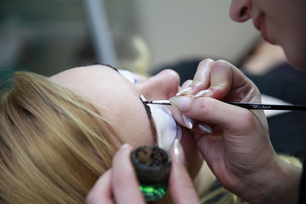 Stylistka brwi precyzyjnie aplikuje hennę na brwi klientki leżącej na fotelu, z widocznymi wacikami ochronnymi pod oczami.