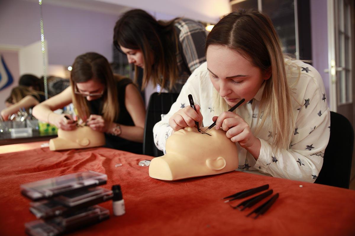 Kurs przedłużania rzęs: Uczestniczki ćwiczą aplikację rzęs na manekinie głowy z bliska, widoczne pęsety i opakowania rzęs na czerwonym stole.