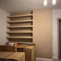 MIELCZAREK CORPORATION - Pomalowana ściana z wbudowanymi półkami w kolorze beżowym, częściowo zasłonięta stertą kartonów na podłodze z panelami, dwie wiszące lampy w tle.