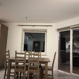 MIELCZAREK CORPORATION - Jasne wnętrze jadalni z drewnianym stołem i krzesłami, widok na okno z roletami i przesuwne drzwi balkonowe, na ścianie widoczny ślad po taśmie malarskiej.