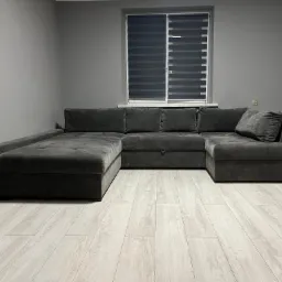 Szary, welurowy narożnik z funkcją spania na jasnej podłodze z paneli, ogrzewany przez elektryczny grzejnik halogenowy, szare ściany, okno z żaluzjami dzień-noc.