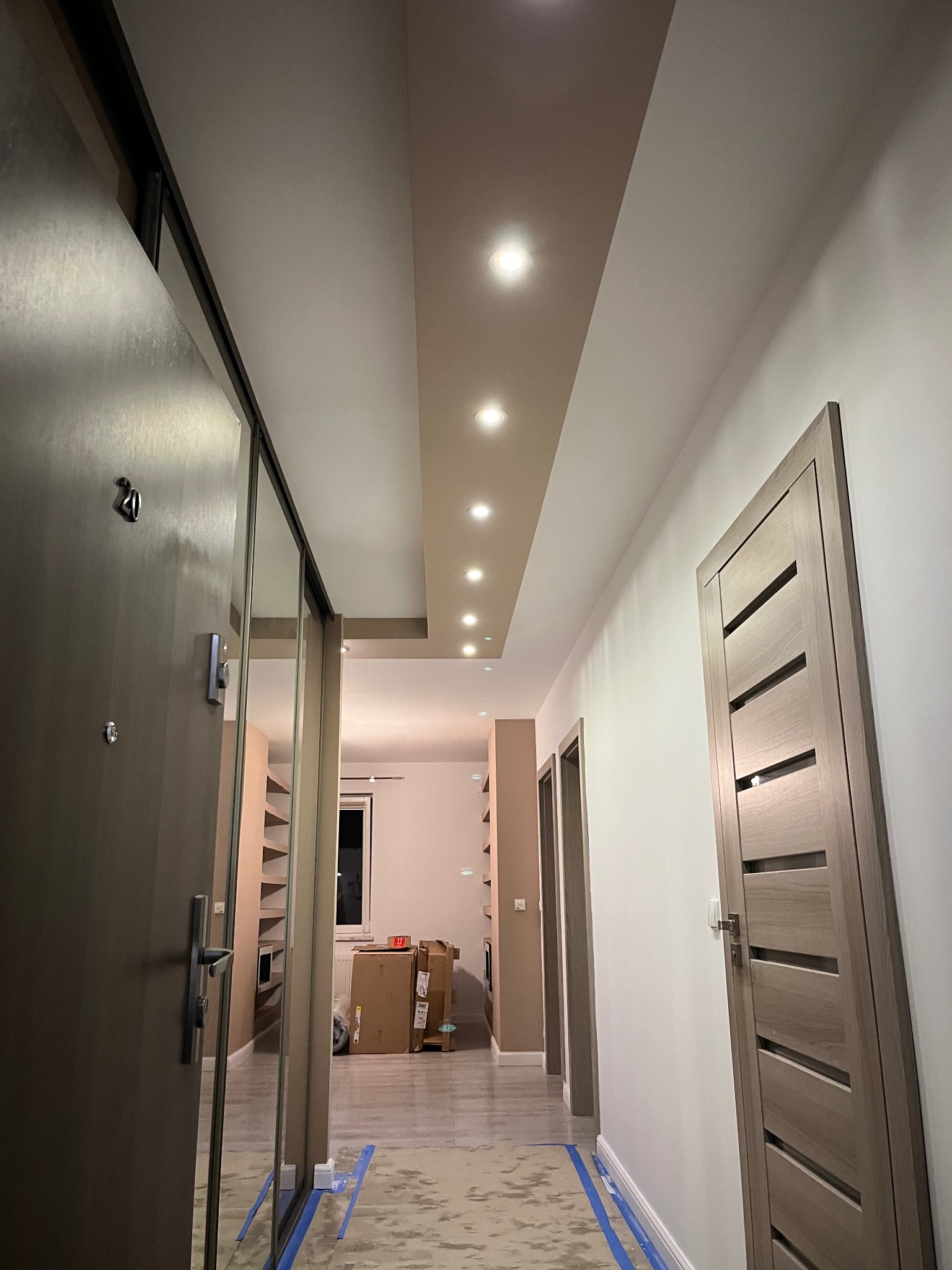 Wnętrze korytarza w trakcie prac wykończeniowych. Widoczna nowa, szara, przesuwna szafa z lustrem, drzwi wewnętrzne w kolorze jasnego drewna, sufit z oświetleniem punktowym i zabezpieczona folią...
