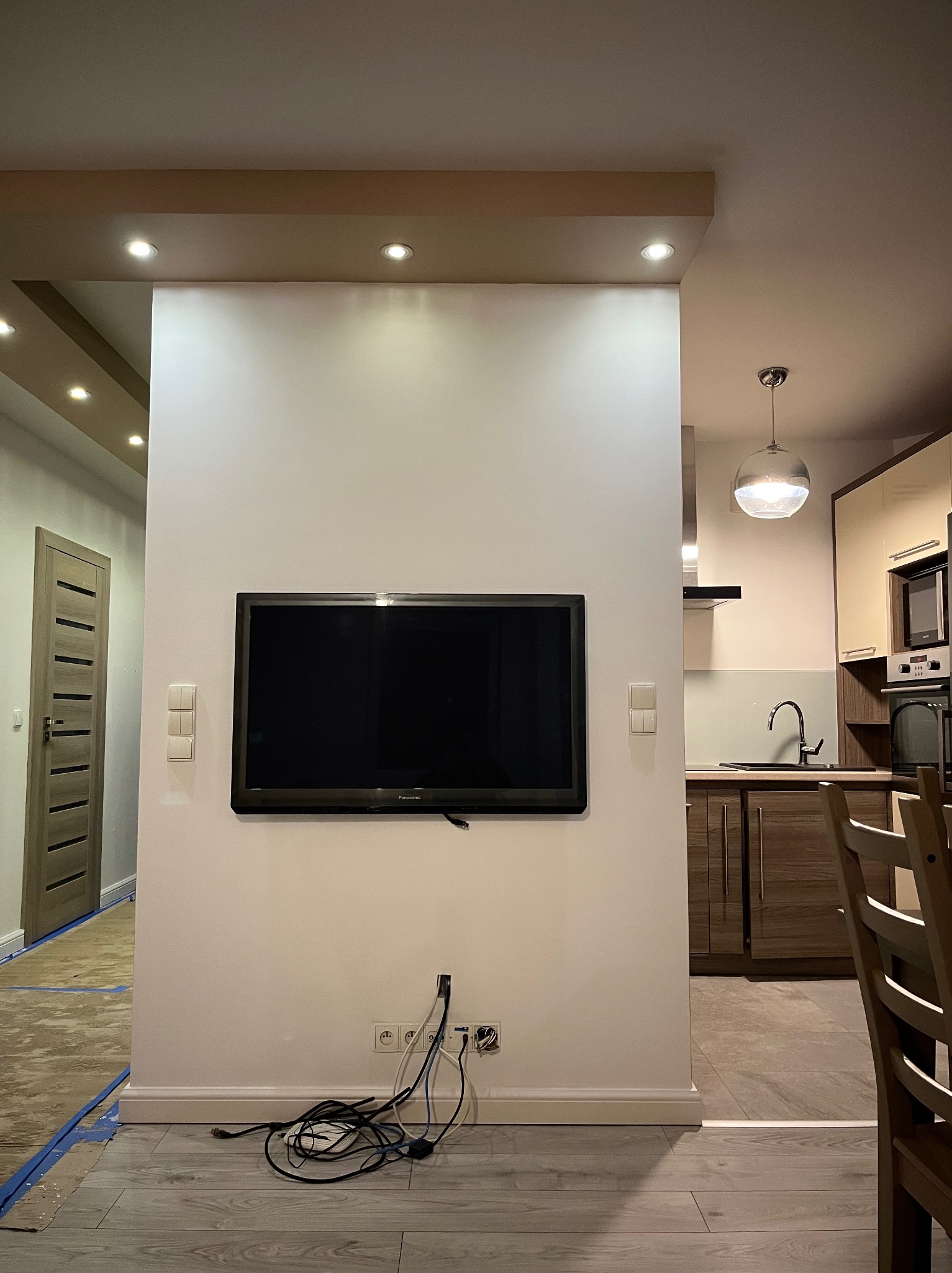 Ściana działowa z zamontowanym telewizorem w nowocześnie urządzonym wnętrzu, widoczna kuchnia w tle, kable zwisające spod telewizora, podłoga w trakcie prac wykończeniowych zabezpieczona taśmą.